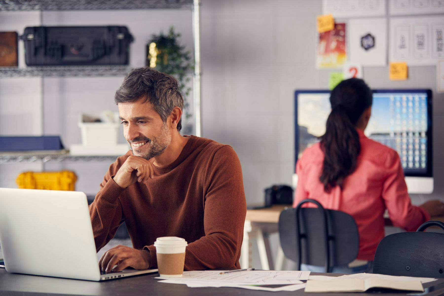 man sitting at a desk looking at computer smiling with woman in background working